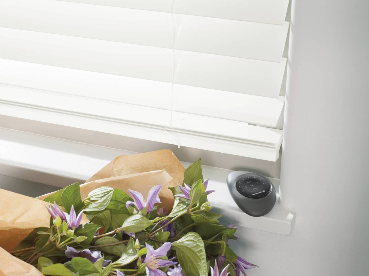 Hunter Douglas PowerView® Pebble® Remote below motorized blinds and beside a purple flower bouquet at House To A Home Interiors near Springfield, IL